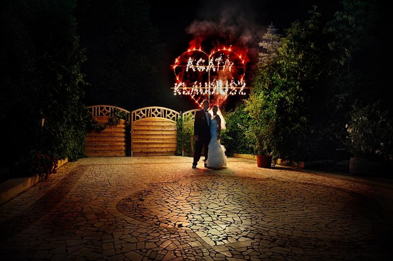 Para młoda stoi na tle płonącego serca z fajerwerków z ich imionami na tle drewnianej bramy i zieleni.