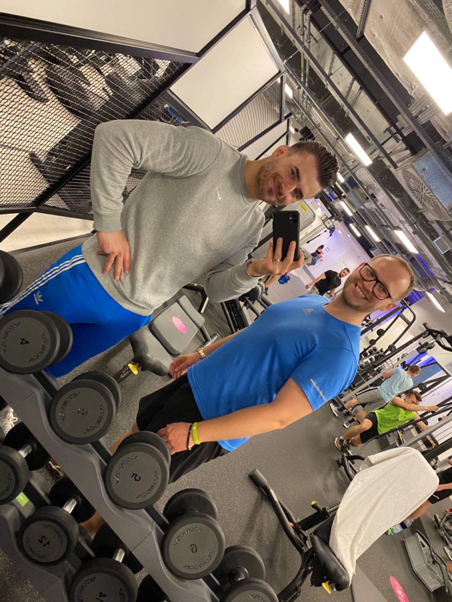 Dwóch uśmiechniętych mężczyzn robi selfie w siłowni, na pierwszym planie stojak z hantlami Technogym, w tle ćwiczący ludzie.