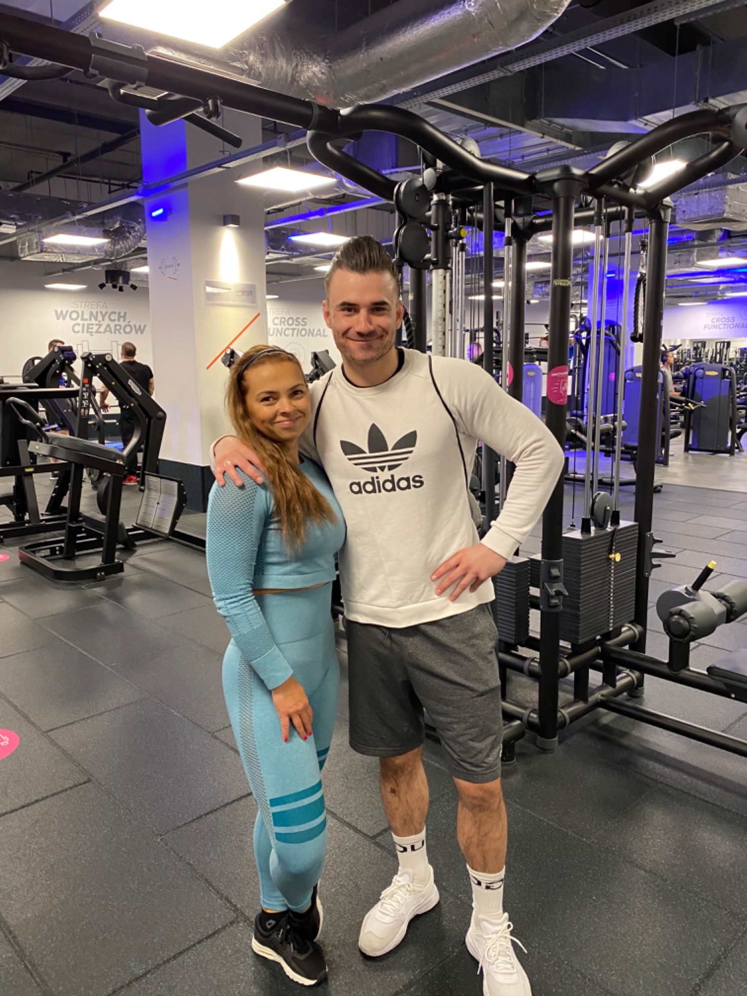 Uśmiechnięta kobieta w błękitnym sportowym stroju i mężczyzna w białej bluzie Adidas pozują na tle maszyn w siłowni.