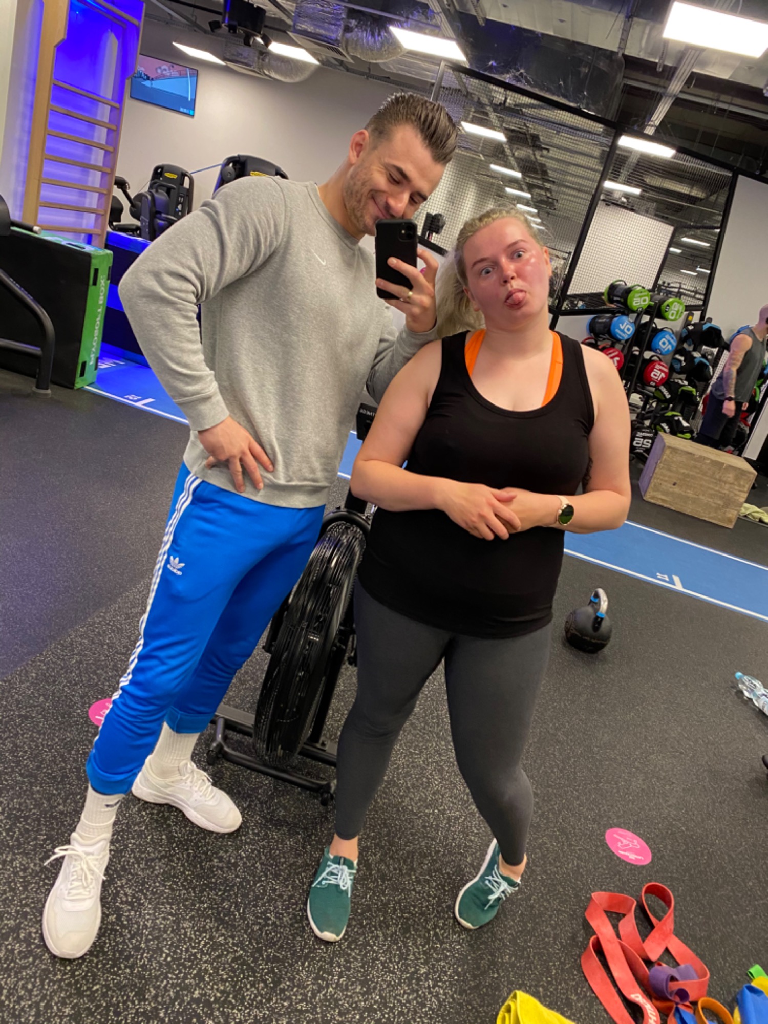 Mężczyzna w szarym swetrze i niebieskich spodniach Adidas robi selfie z kobietą w czarnym topie na siłowni, kobieta wystawia język.