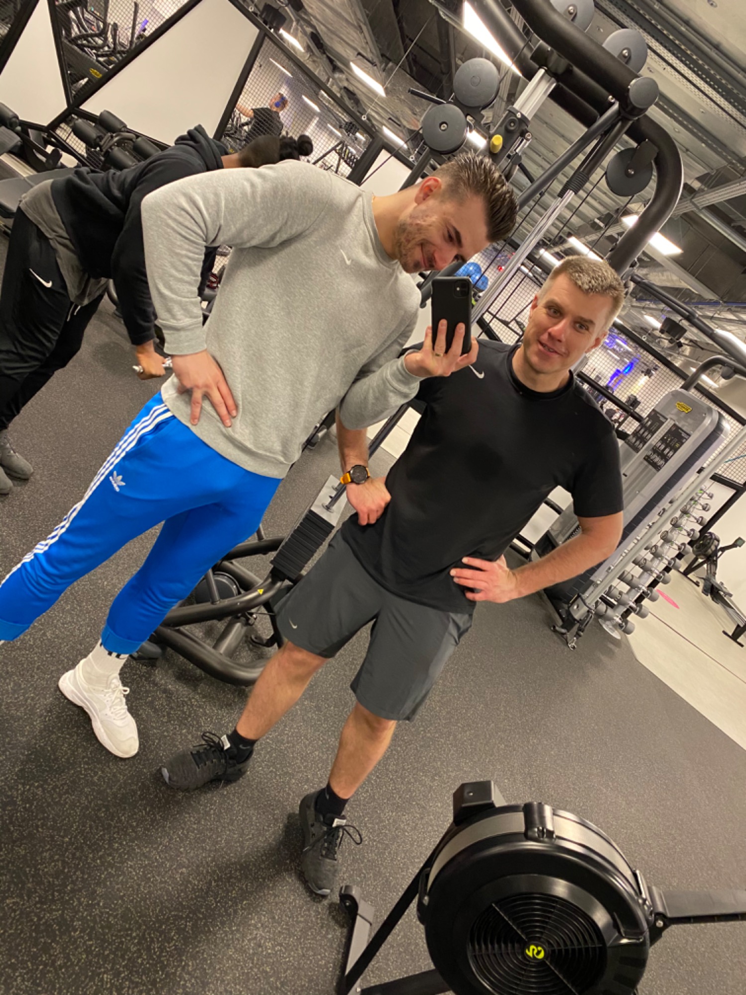 Dwóch mężczyzn w sportowych strojach pozuje do selfie w nowoczesnej siłowni, widoczny sprzęt do ćwiczeń i osoby trenujące w tle.