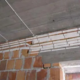 Instalacja elektryczna w trakcie montażu: białe kable ułożone wzdłuż ceglanej ściany pod surowym betonowym sufitem. Widoczne otwory w ścianie i prowadzone przewody.