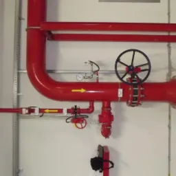 System rur przeciwpożarowych w kolorze czerwonym z zaworami, manometrem i czarnym kołem sterującym, zamontowany na ścianie z widocznymi elementami mocującymi.