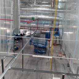 NAPTECH Dariusz Napieracz - Widok z góry na wnętrze hali przemysłowej z instalacjami pod sufitem, z widocznymi kanałami wentylacyjnymi, rurami, trasami kablowymi i podnośnikiem koszowym na posadzce, zabezpieczonej folią...