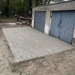 Świeżo ułożona, szara kostka brukowa przed dwustanowiskowym garażem z szarymi drzwiami, widoczne pozostałości piasku i ziemi po pracach.