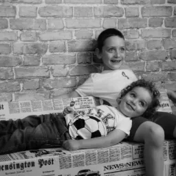 Dw&oacute;ch chłopc&oacute;w w czarno-białym portrecie: starszy opiera się o ceglaną ścianę, młodszy leży na gazecie, obaj uśmiechnięci.