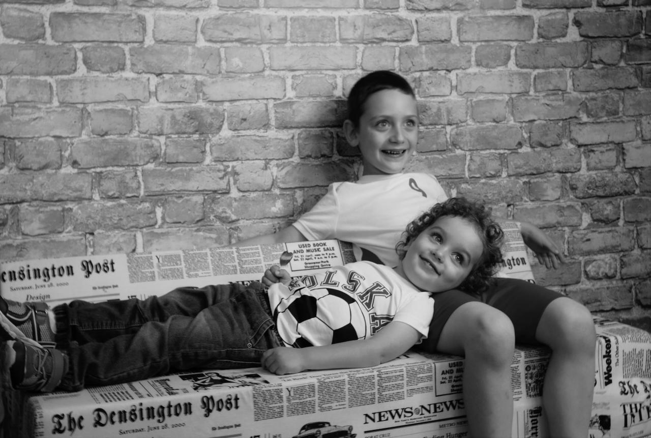 Dwóch chłopców w czarno-białym portrecie: starszy opiera się o ceglaną ścianę, młodszy leży na gazecie, obaj uśmiechnięci.