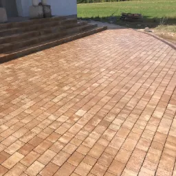 Nowo ułożony, beżowo-brązowy plac z kostki, prowadzący do schodów wejściowych budynku, z widocznymi paletami w tle.