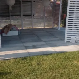 Wyłożona szarymi płytami strefa relaksu z białym ażurowym zadaszeniem, widoczne dekoracyjne lampiony i trawnik.