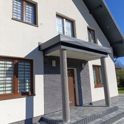 Firma TIMBER Artur Terpiłowski - Nowoczesny dom jednorodzinny z szarą elewacją imitującą kamień przy wejściu, zadaszeniem wspartym na okrągłych kolumnach i schodami z kostki brukowej.