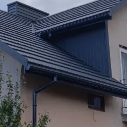 Nowy, grafitowy dach z blachodachówki modułowej, rynny w tym samym kolorze, wykończony komin z blachy, widok z dołu na fragment elewacji budynku.