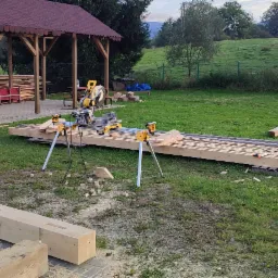 Przygotowanie drewnianych elementów konstrukcyjnych na trawie, widoczna piła stołowa i długa belka z wyżłobieniami, w tle zadaszenie i zielony krajobraz.