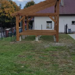 Drewniana konstrukcja altany z prostym dachem na betonowych podstawach, widoczna na tle trawnika i budynku z czerwonym dachem.