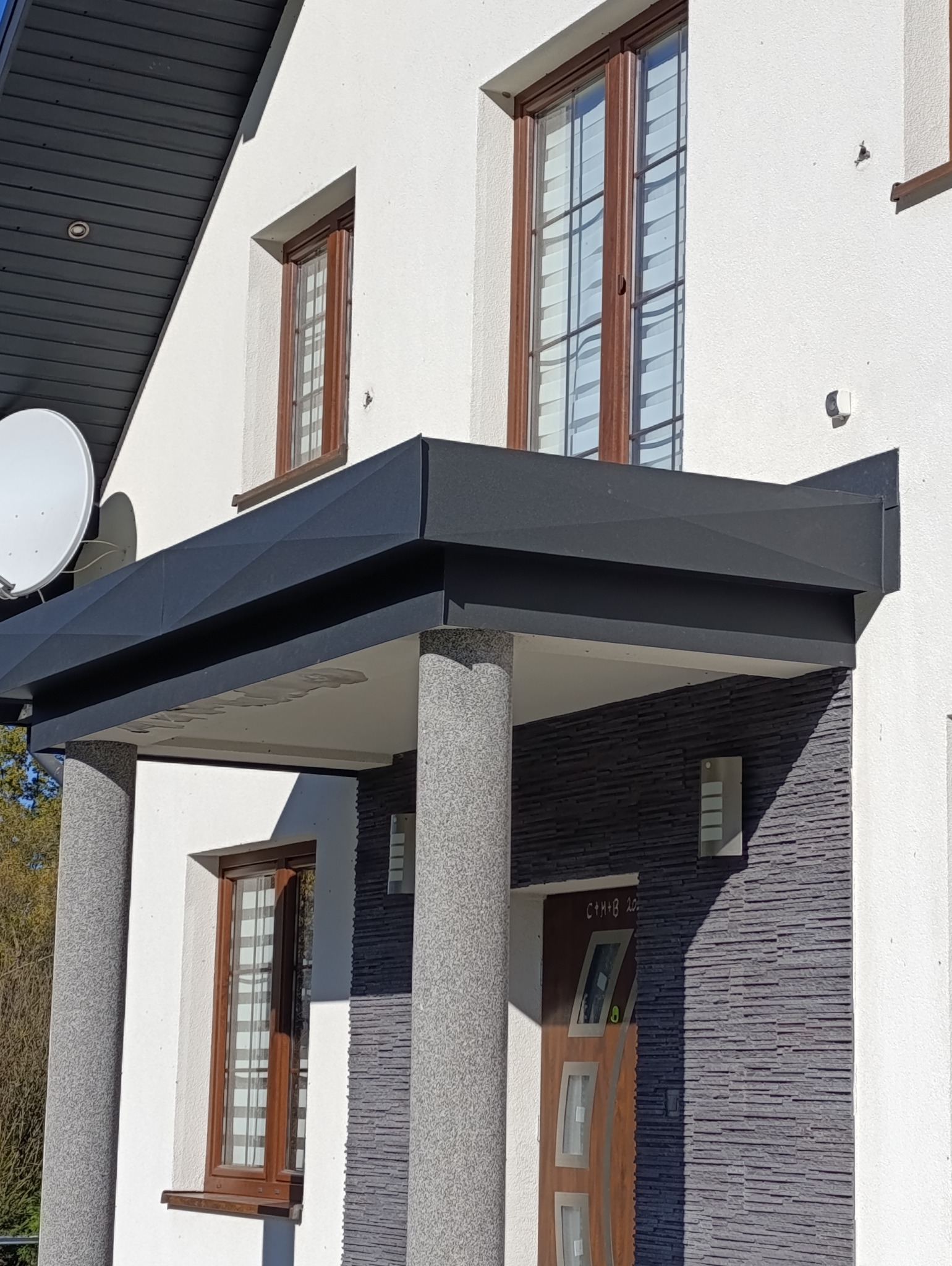 Nowoczesne zadaszenie nad wejściem do domu z białymi ścianami, kamienną elewacją i kolumnami o fakturze lastryko, widoczna antena satelitarna.