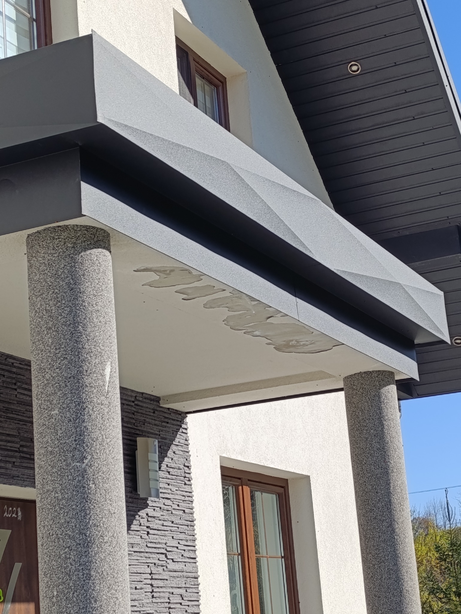 Nowoczesny daszek nad wejściem do domu z szarej blachy trapezowej, wsparty na dwóch okrągłych, kamiennych kolumnach, z widocznymi śladami po kleju montażowym na spodniej stronie daszku.