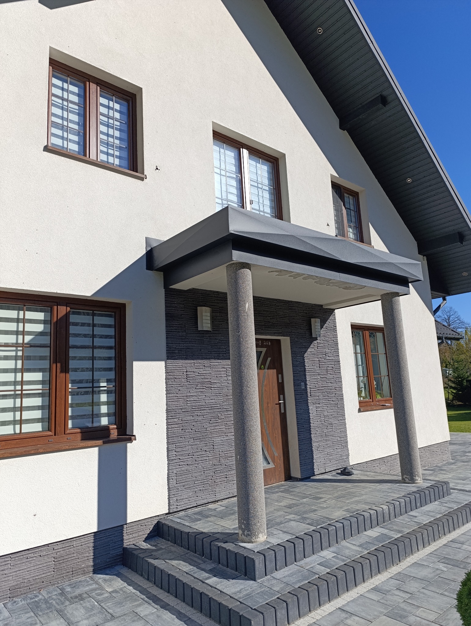 Nowoczesny dom jednorodzinny z szarą elewacją imitującą kamień przy wejściu, zadaszeniem wspartym na okrągłych kolumnach i schodami z kostki brukowej.