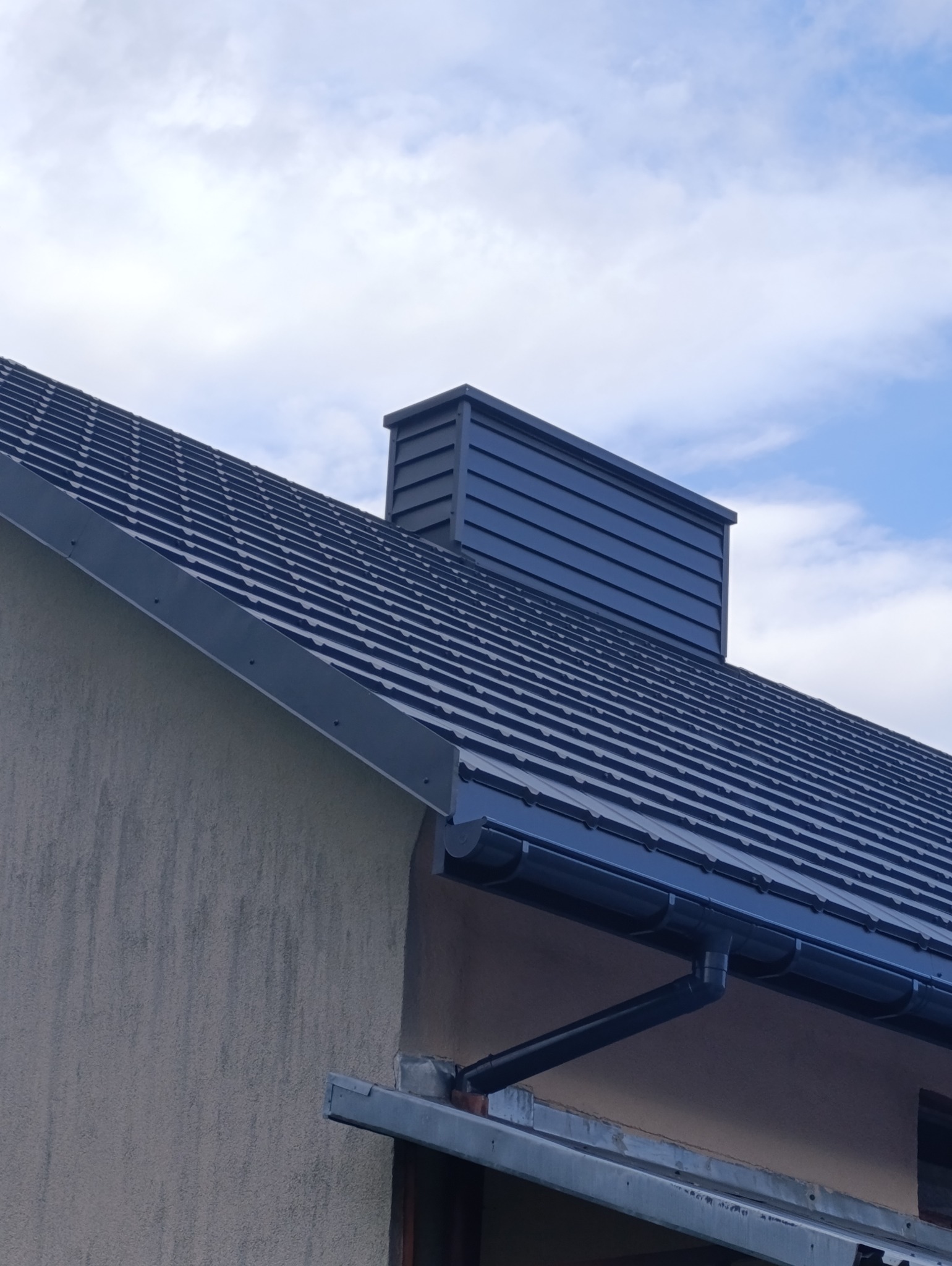 Nowy, grafitowy dach z blachy trapezowej, z widocznym kominem i systemem rynnowym, na tle błękitnego nieba z chmurami.