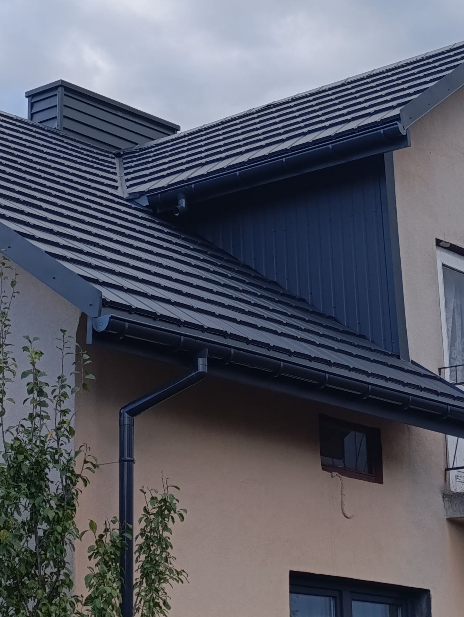 Nowy, grafitowy dach z blachodachówki modułowej, rynny w tym samym kolorze, wykończony komin z blachy, widok z dołu na fragment elewacji budynku.