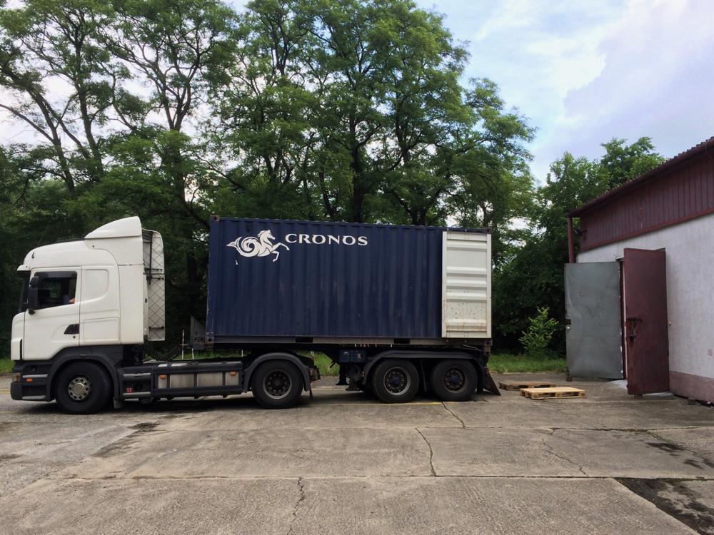 Ciężarówka z niebieskim kontenerem firmy Cronos na placu przed budynkiem z otwartymi drzwiami, w tle drzewa.