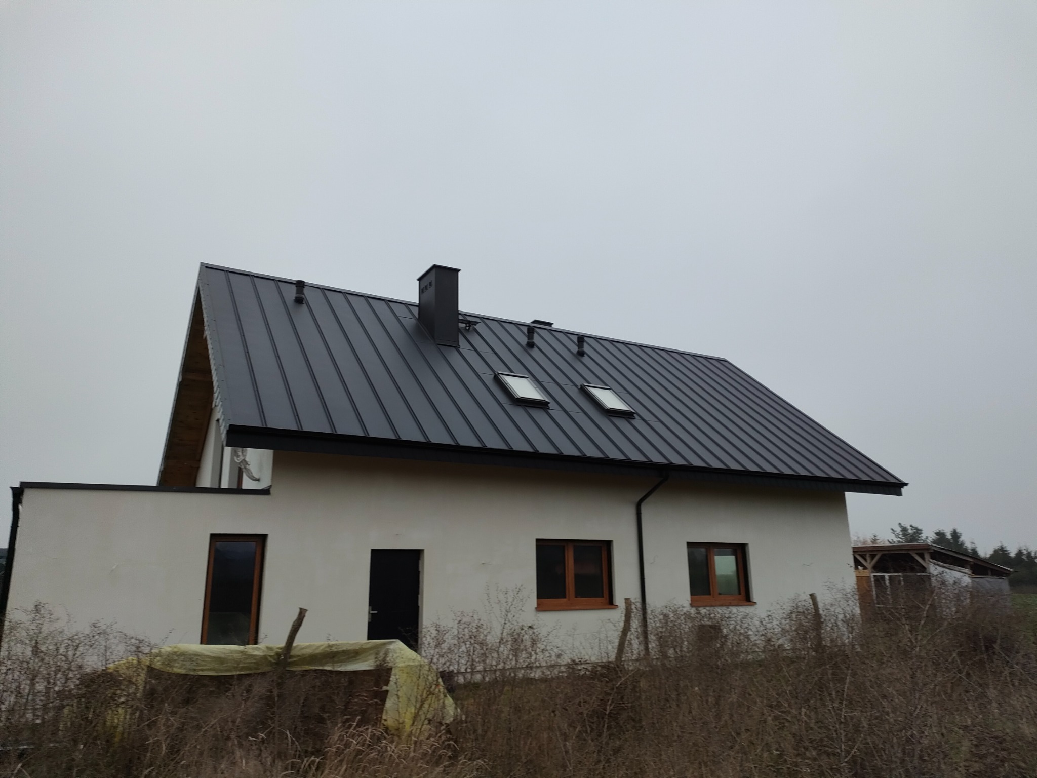 Dom jednorodzinny z ciemnoszarym dachem z blachy na rąbek stojący, widoczne okna dachowe i komin, elewacja w trakcie wykańczania, teren wokół domu niezagospodarowany.