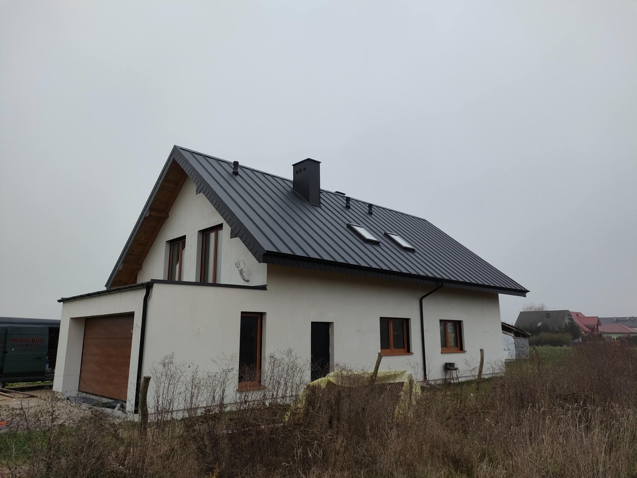 Nowoczesny dom jednorodzinny z grafitowym dachem z blachy na rąbek stojący, białymi ścianami i drewnianą stolarką okienną, widziany z poziomu gruntu w pochmurny dzień. W tle widać zabudowę...