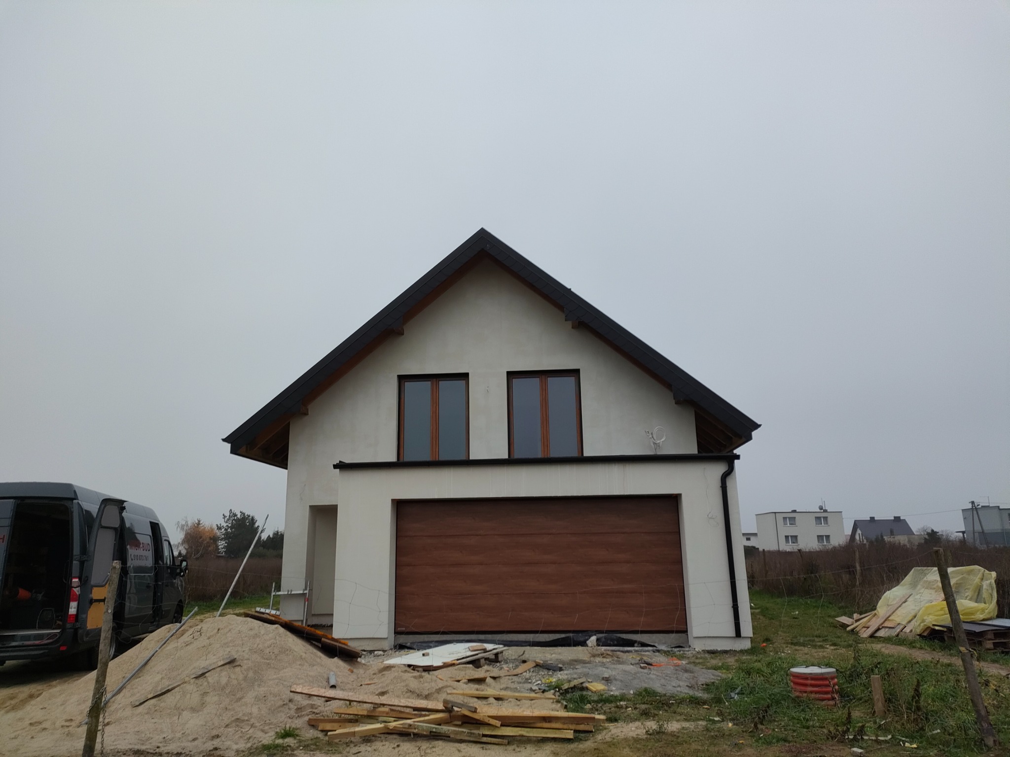 Nowo wybudowany dom jednorodzinny z brązową bramą garażową i białymi ścianami zewnętrznymi, widoczny stos piasku i resztki materiałów budowlanych, szara pogoda.