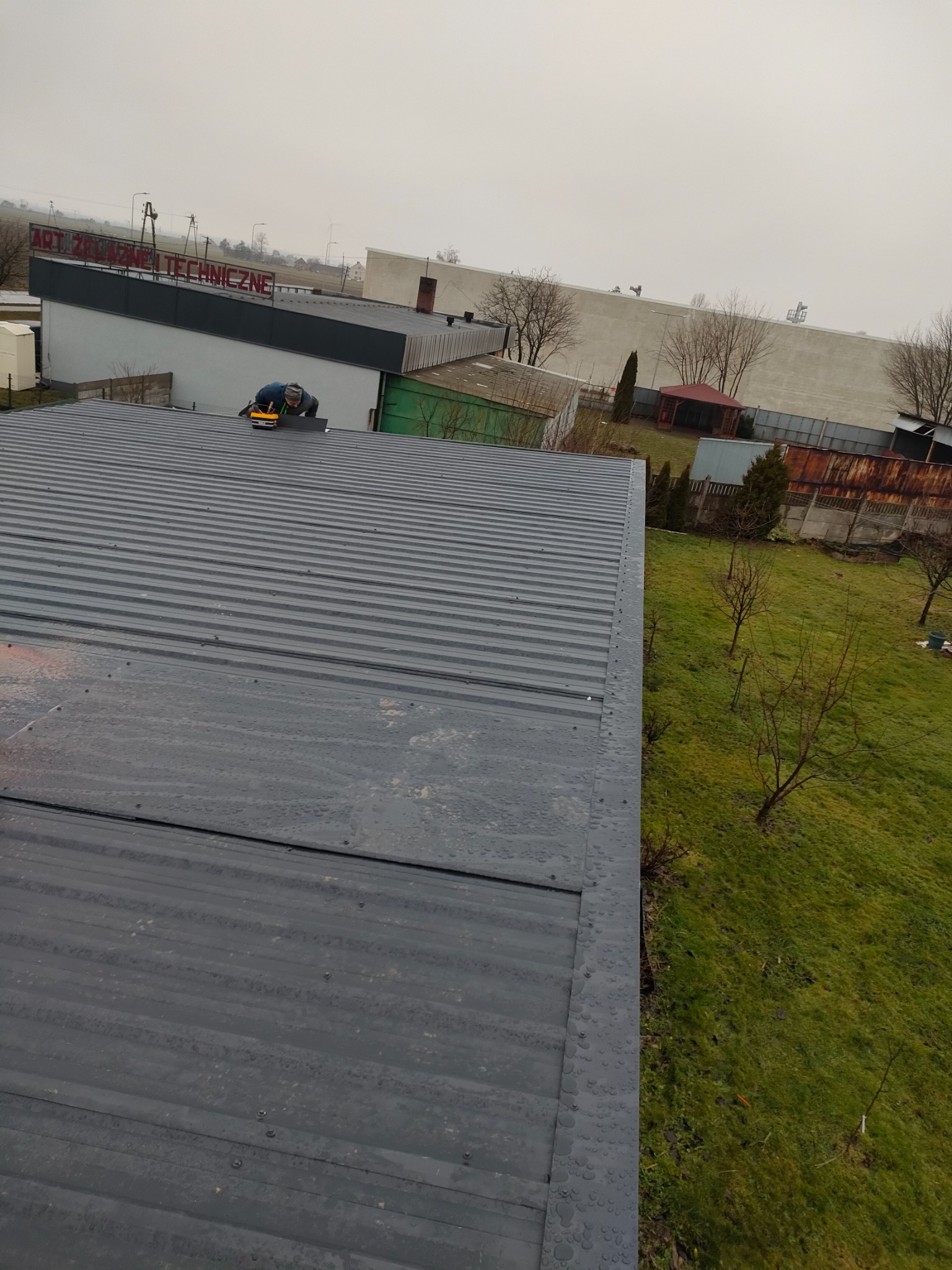 Pracownik w trakcie montażu blachy trapezowej na dachu budynku gospodarczego, widok z góry na szare poszycie z widocznymi wkrętami, w tle budynki przemysłowe i zieleń.