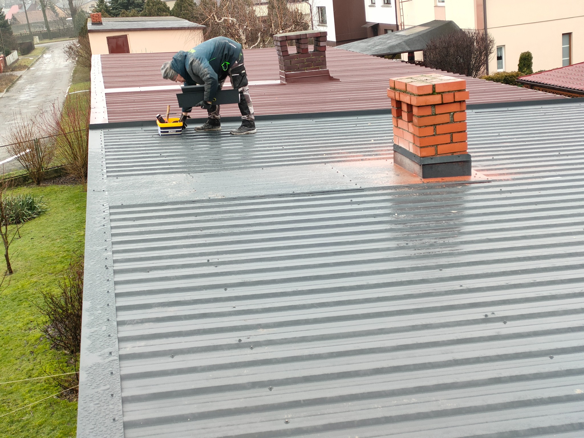 Monter w roboczym ubraniu, w trakcie instalacji elementu na szarym dachu z blachy trapezowej, obok komin z cegły i skrzynka narzędziowa.