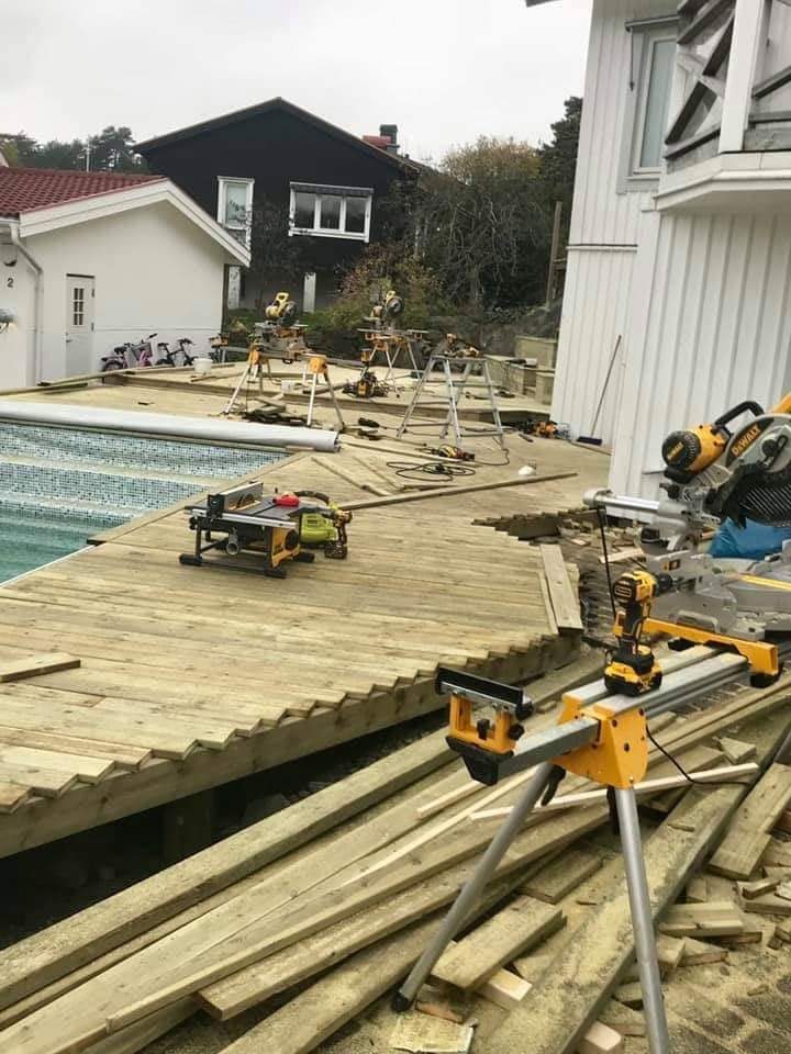 Budowa drewnianego tarasu przy basenie: widoczne deski, narzędzia do cięcia i montażu, częściowo ukończona konstrukcja z charakterystycznym ząbkowanym wykończeniem krawędzi, w tle domy i drabiny.