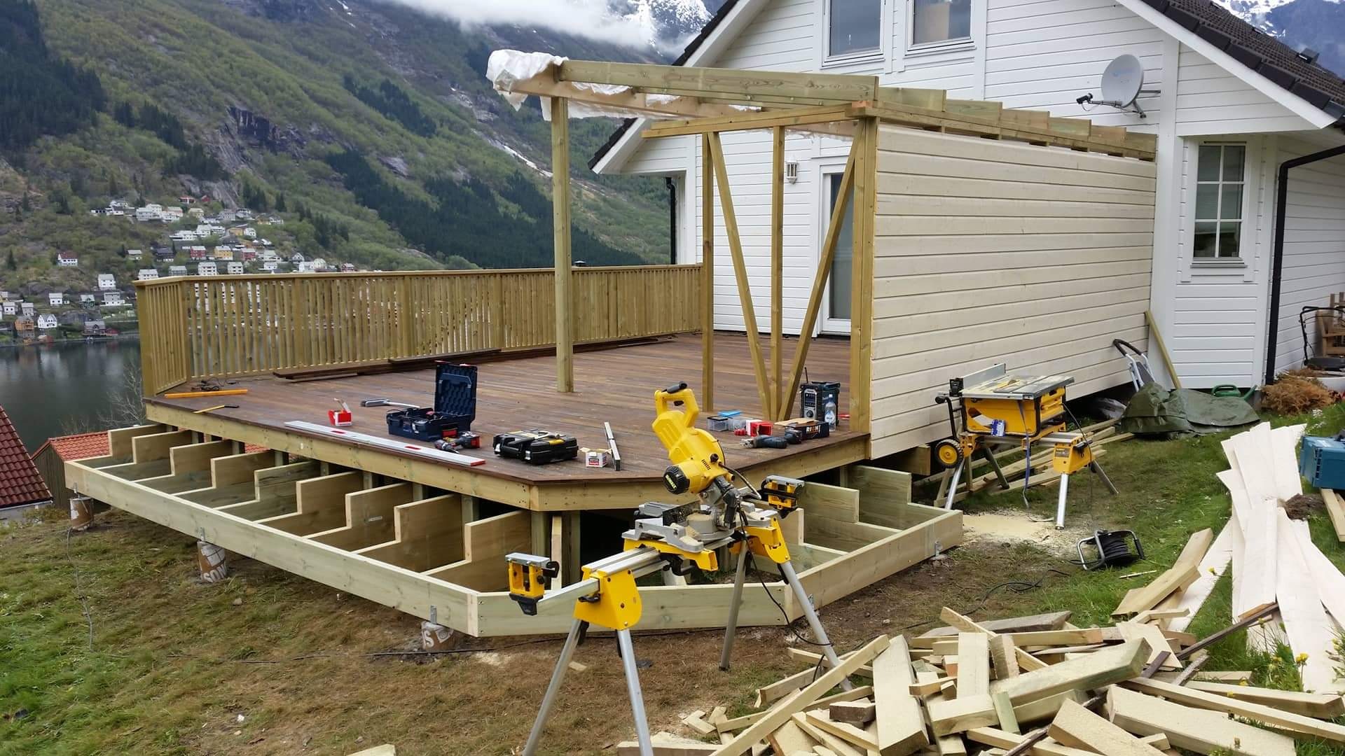Budowa tarasu drewnianego z balustradą przy domu, widoczne narzędzia, konstrukcja nośna, tło górskie z jeziorem.