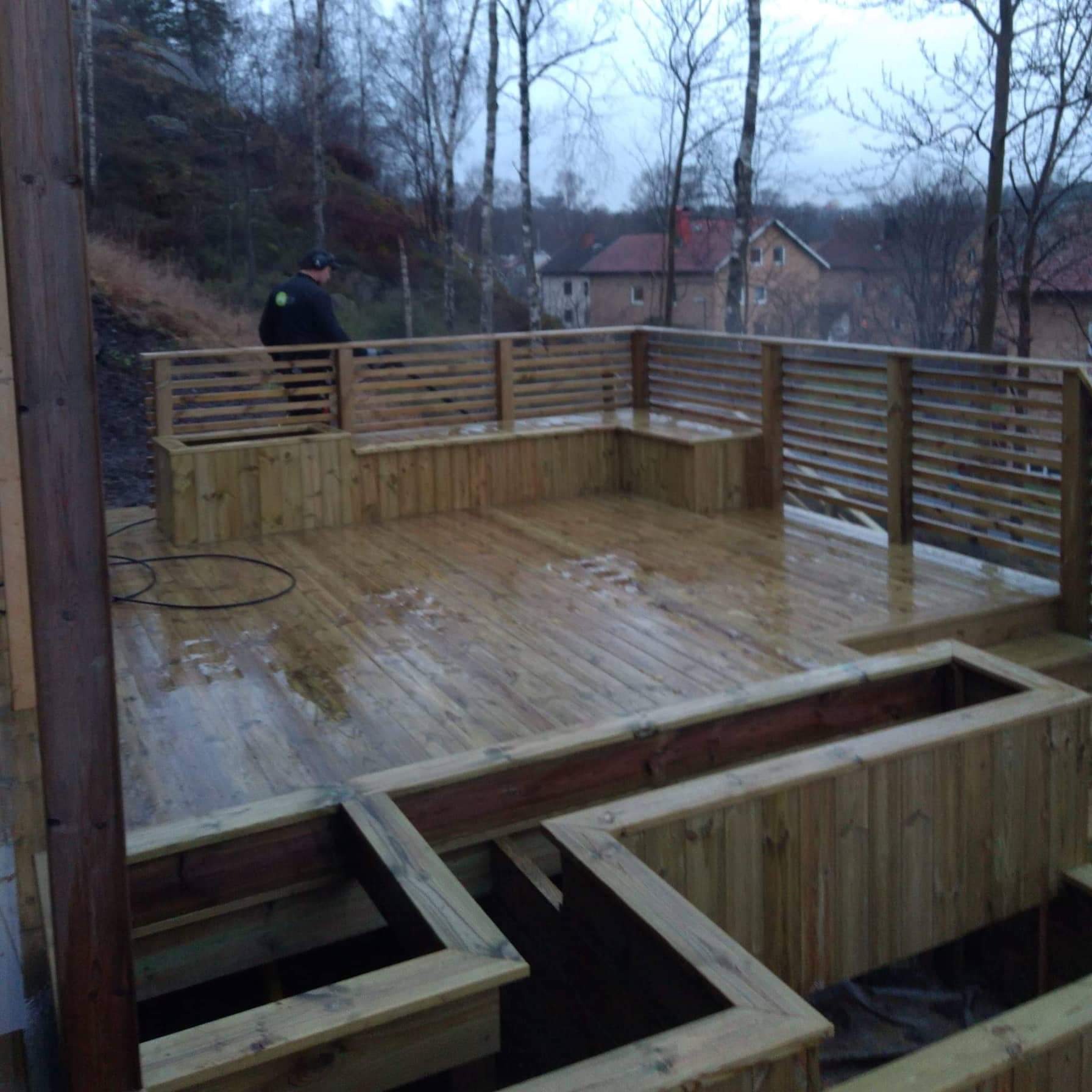 Nowy drewniany taras z wbudowanymi siedziskami i donicami, widok na okoliczne domy i drzewa w pochmurny dzień.