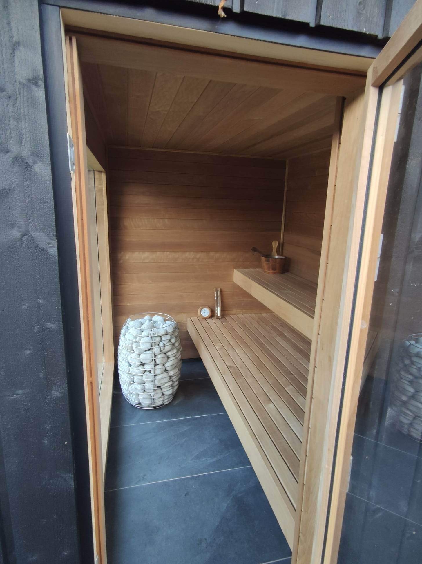 Wnętrze sauny z drewnianymi ławkami, wiaderkiem i nabierką, termometrem oraz koszem z białymi kamieniami, widoczne przez otwarte drzwi z drewnianą ramą i szklanym panelem.