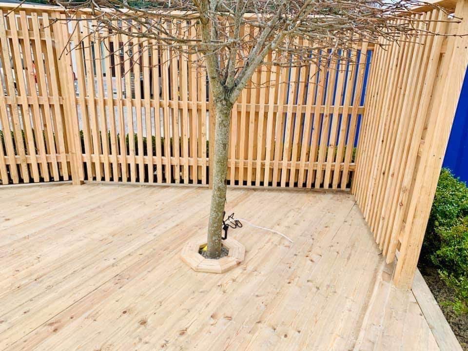 Nowoczesny taras z jasnego drewna otoczony płotem z pionowych desek, z drzewem rosnącym pośrodku i osłoniętym drewnianą obudową u podstawy.