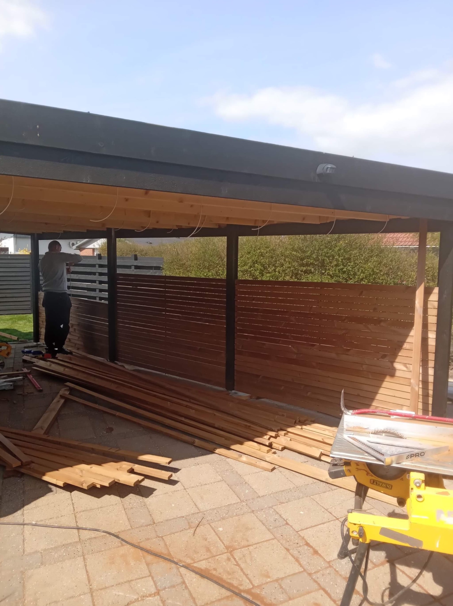 Budowa drewnianej konstrukcji z zadaszeniem, widoczny pracownik przytwierdzający deski do ściany, na pierwszym planie leżą docięte elementy oraz piła stołowa.