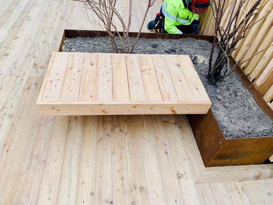 Jasny drewniany taras z wbudowaną rabatą na rośliny w metalowej obudowie, częściowo zasłonięty pracownikiem w odblaskowej kurtce.