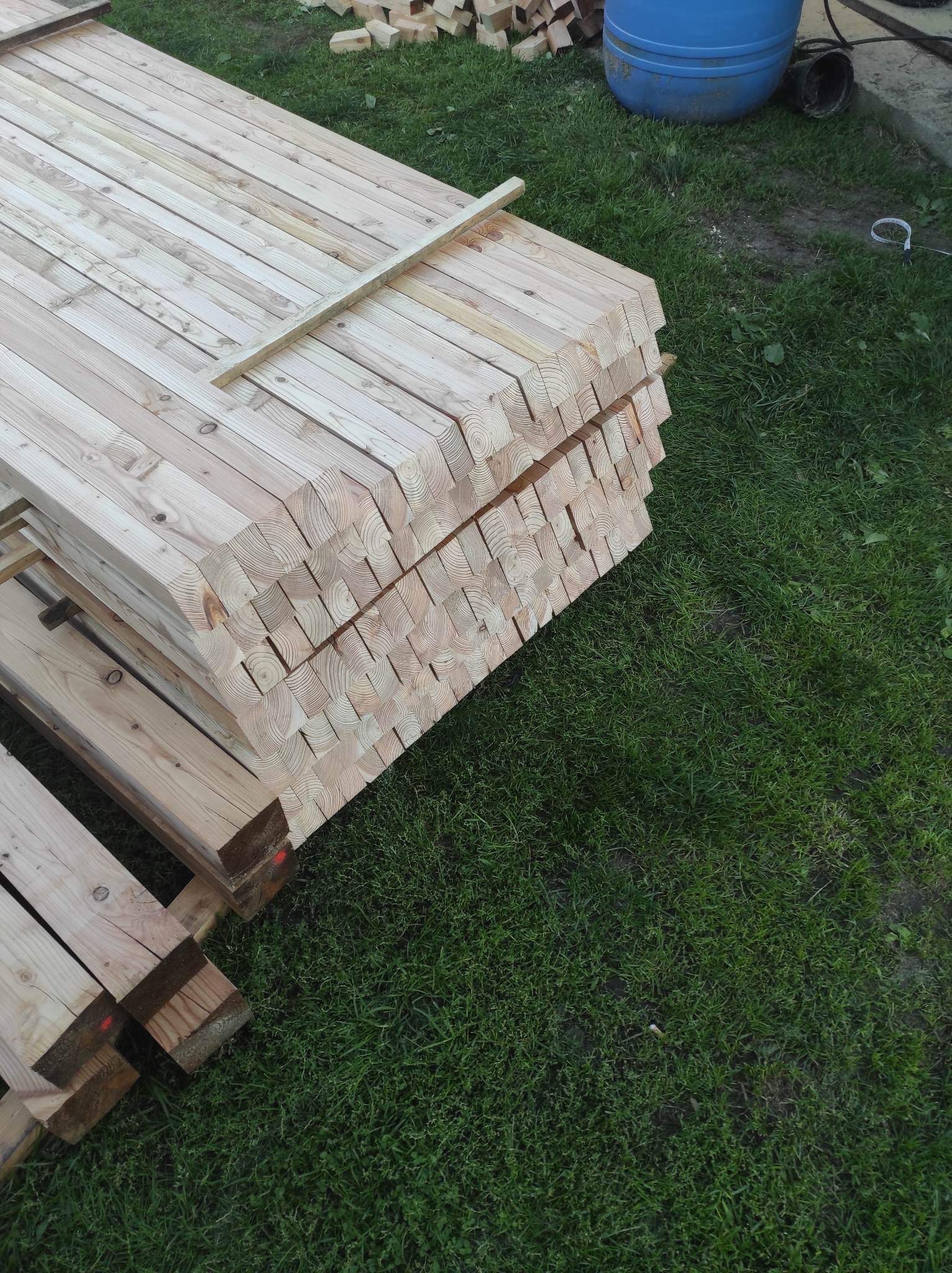 Stos świeżo obrobionych belek drewnianych ułożonych na palecie na trawie, widoczne słoje drewna na przekroju, przygotowane do dalszej obróbki lub montażu.