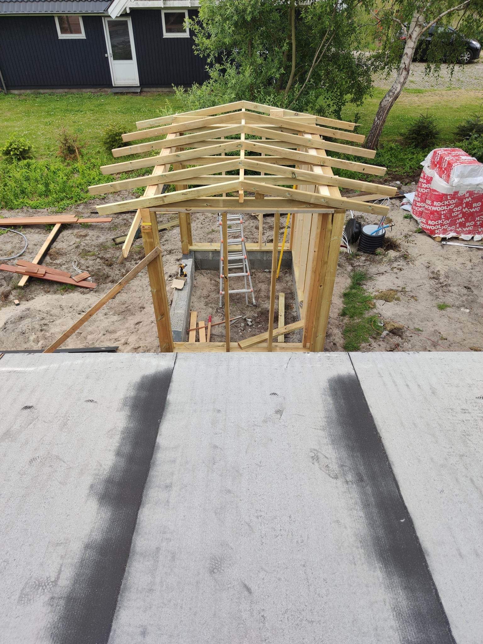 Budowa drewnianej konstrukcji z dachem w ogrodzie, widok z góry na szkielet z belek, w tle budynek mieszkalny i materiały izolacyjne.