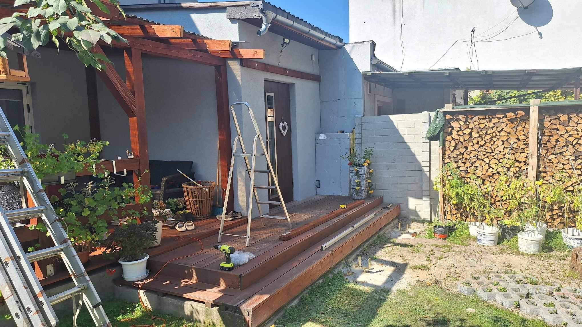 Budowa tarasu drewnianego: deski tarasowe w kolorze brązowym, drabina, wkrętarka akumulatorowa i elementy konstrukcyjne na tle domu z drewnianymi drzwiami i altany.