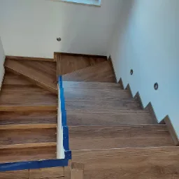 Schody wewnętrzne wyłożone płytkami imitującymi drewno, zabezpieczone niebieską taśmą malarską na krawędziach, z widocznym oknem dachowym i punktowym oświetleniem ściennym.