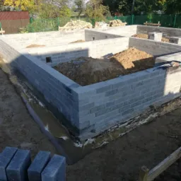 Wznoszenie fundamentów z bloczków betonowych, widoczne wykopane rowy, izolacja przeciwwilgociowa oraz pryzmy ziemi wewnątrz zarysu fundamentów.