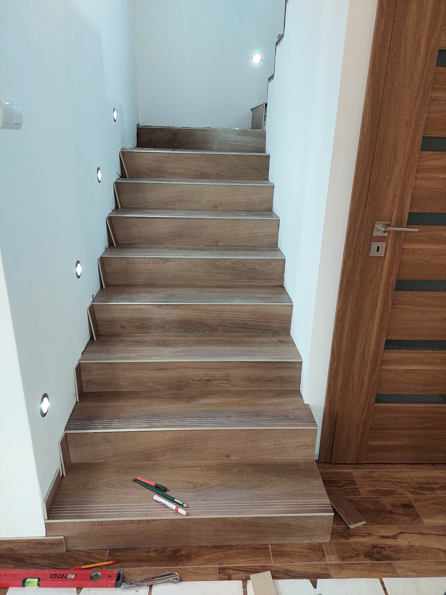 Montaż drewnopodobnych paneli na schodach z podświetleniem LED w ścianie, widoczne narzędzia: poziomica, ołówki, nóż do tapet.