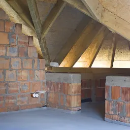 Wnętrze poddasza w trakcie budowy: ściany z czerwonej cegły, surowa wylewka podłogowa, widoczne drewniane elementy konstrukcji dachu i instalacja elektryczna z białymi gniazdkami.