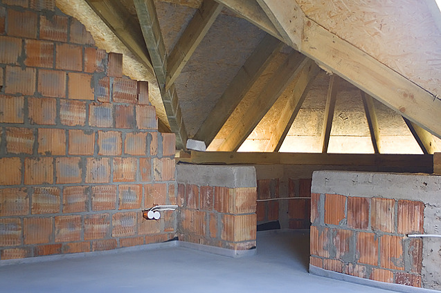 Wnętrze poddasza w trakcie budowy: ściany z czerwonej cegły, surowa wylewka podłogowa, widoczne drewniane elementy konstrukcji dachu i instalacja elektryczna z białymi gniazdkami.