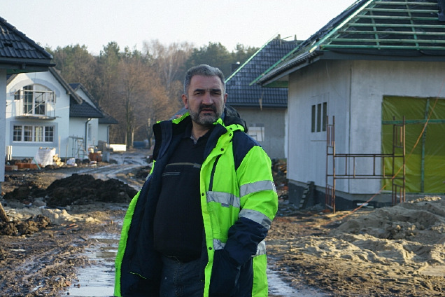 Mężczyzna w odblaskowej kurtce na tle budowy domów jednorodzinnych w Otwocku, widoczny teren budowy w trakcie prac ziemnych.
