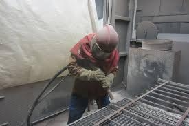 Pracownik w kombinezonie ochronnym i hełmie piaskuje metalową kratę w zamkniętym pomieszczeniu, widoczne przewody i elementy konstrukcyjne.