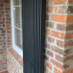 Wysoki, czarny, pionowy grzejnik panelowy zamontowany na ceglanej ścianie obok okna z białymi ramami.