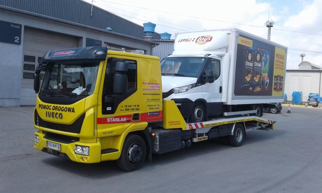 Żółta laweta Iveco z napisem 'Pomoc Drogowa' przewozi białą ciężarówkę z reklamą chipsów Lays na parkingu przed budynkiem.