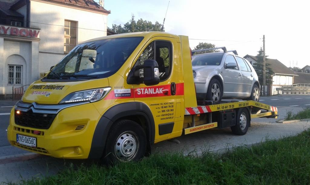 Żółta laweta marki Citroen z logo firmy Stanlak, przewożąca srebrny samochód osobowy na tle budynku miejskiego.