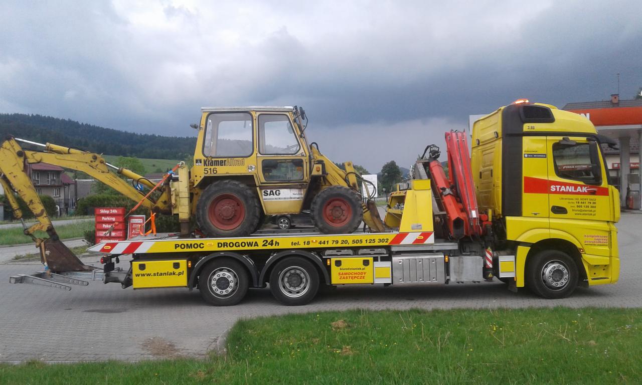 Żółta laweta firmy Stanlak z żółtą koparko-ładowarką KramerAllrad 516 na platformie, zaparkowana na poboczu z widokiem na budynki i zalesione wzgórza w tle.