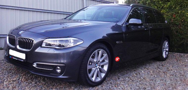 Ciemnoszary samochód BMW kombi zaparkowany na żwirowym podłożu, z widoczną naklejką 'ST' na przednich drzwiach.